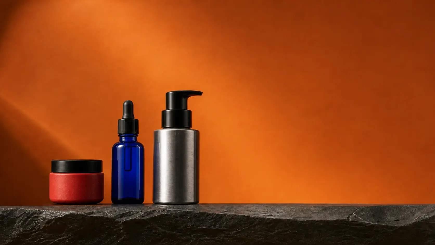 Korallfarbener Tiegel, kobaltblaue Pipettenflasche und silberner Pumpspender vor orangefarbenem Hintergrund